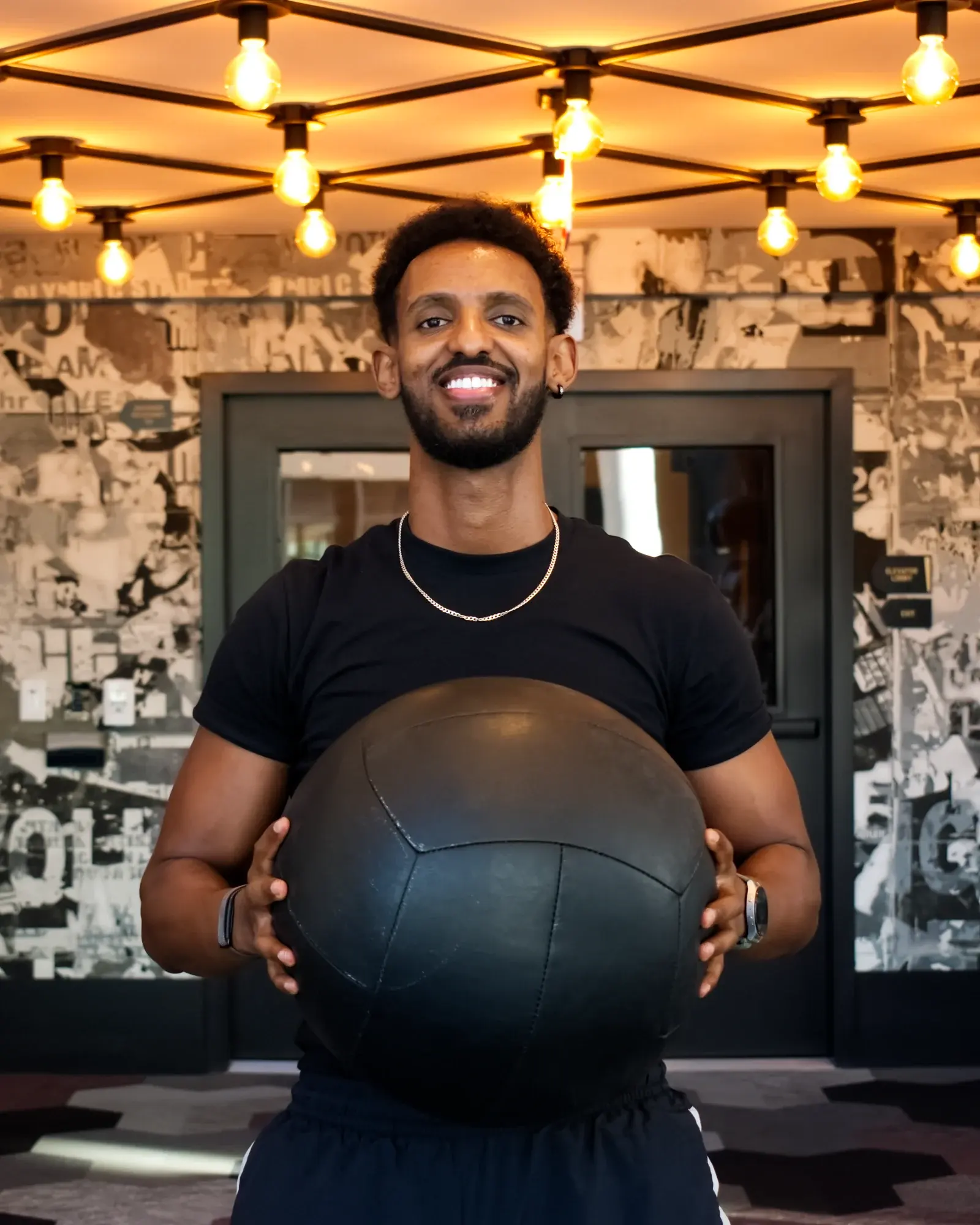 Nathnael Gebre smiling in a gym, holding a black medicine ball in front of him under warm hanging lights.
