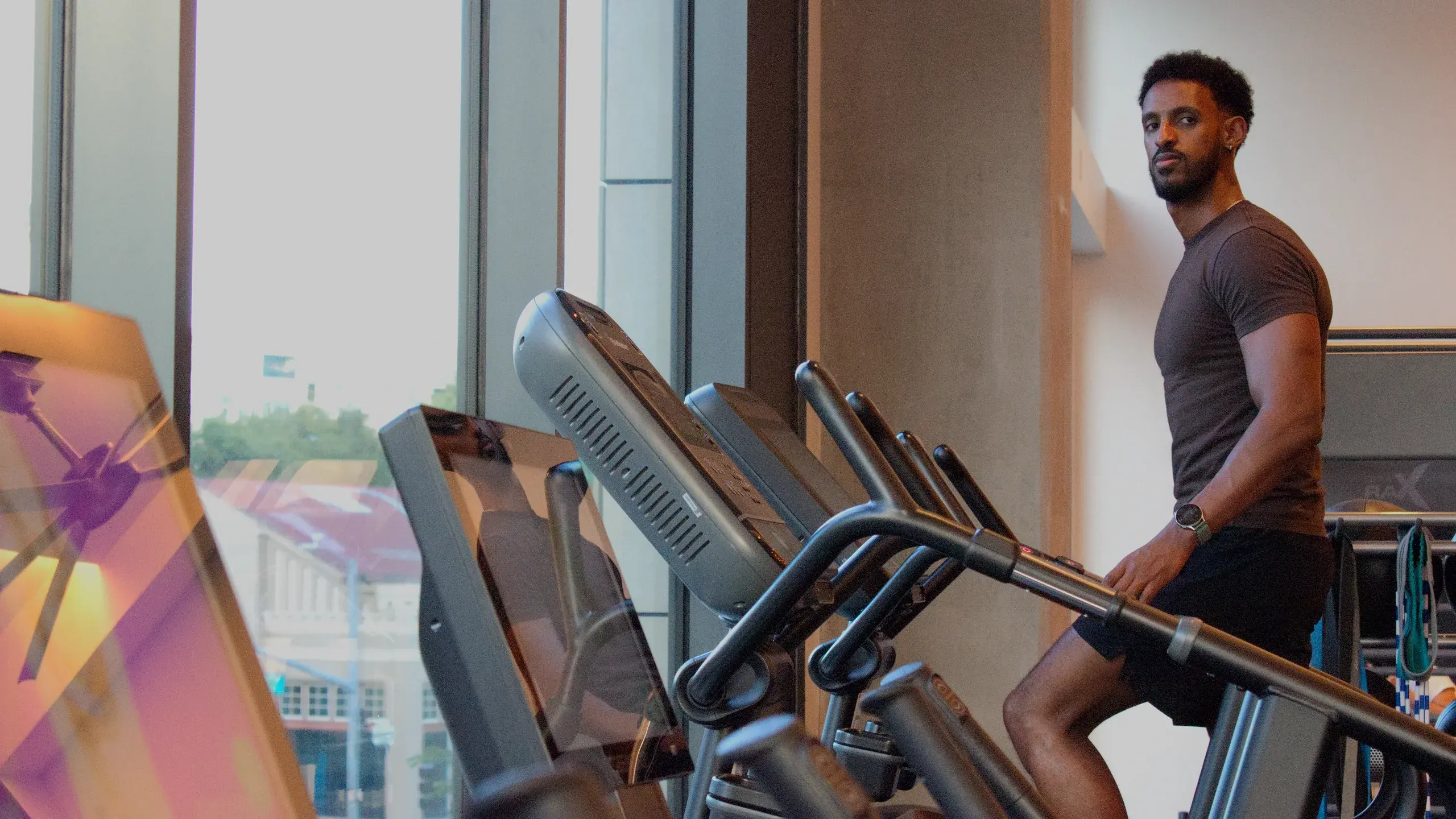 Nathnael Gebre standing beside a row of stair-climber machines next to floor-to-ceiling gym windows.
